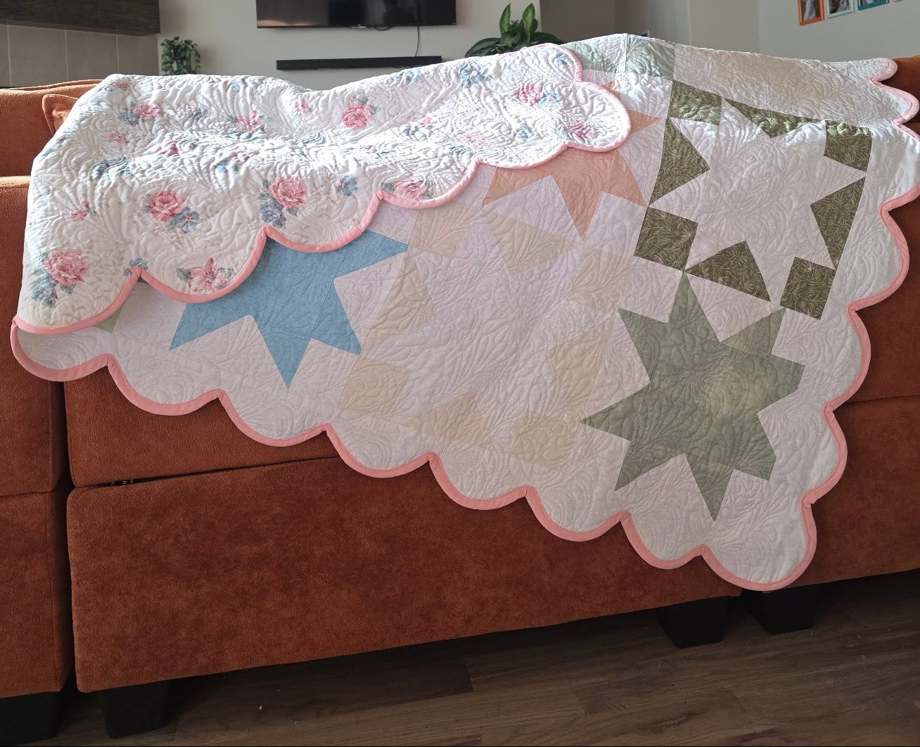 A handmade quilted blanket with a scalloped pink border, featuring a floral pattern on one side and starburst geometric designs on the other, draped over an orange-brown sectional sofa in a living room.